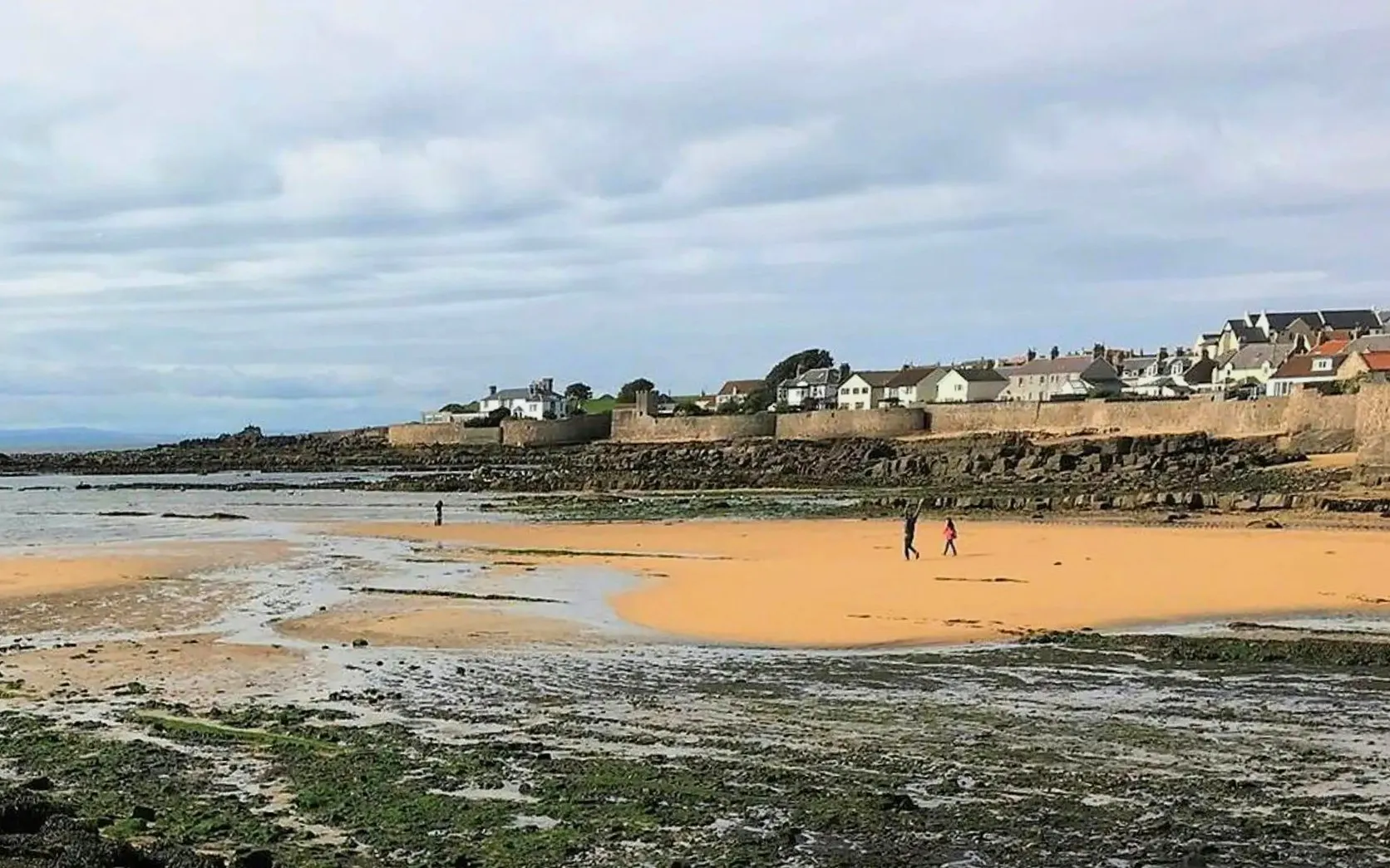 Elie Beach with people walking on sandy shore, coastal village in background.
