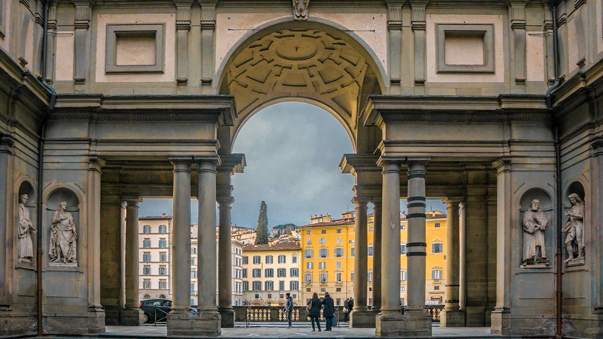Visitors framed by the grand arches of the Uffizi Gallery's entrance, with statues standing sentinel on either side, welcoming art enthusiasts