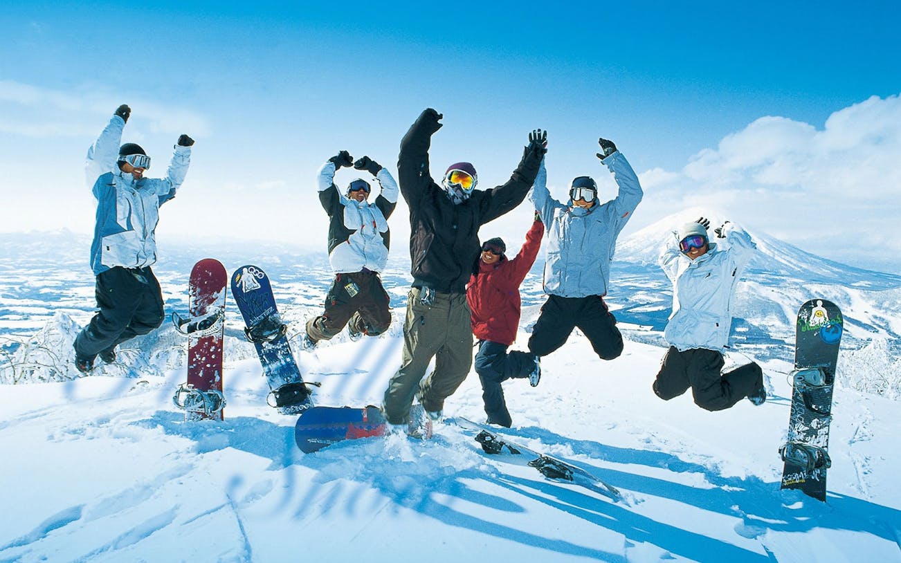 Group of snowboarders jumping on snowy mountain in Niseko, Japan, with snowboards in foreground.