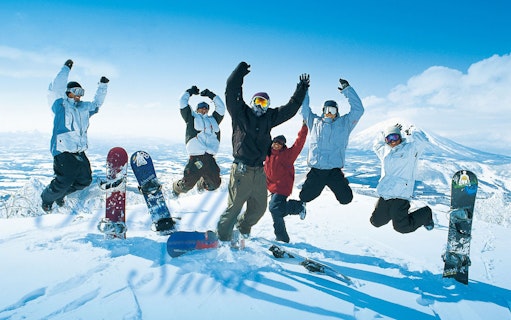Group of snowboarders jumping on snowy mountain in Niseko, Japan, with snowboards in foreground.