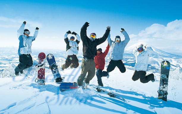 Group of snowboarders jumping on snowy mountain in Niseko, Japan, with snowboards in foreground.
