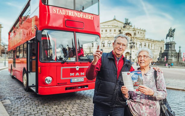 Elderly couple with map in front of red double-decker bus on Dresden city tour.
