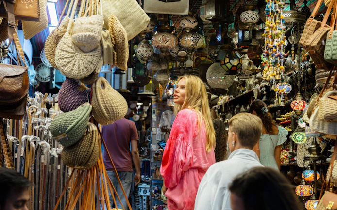 Tourists exploring Muttrah Souk with hanging lanterns and woven bags in Muscat, Oman.