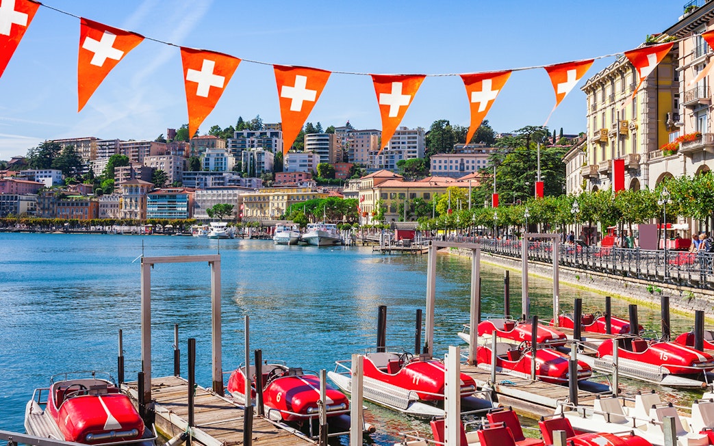 Lake Lugano waterfront with red pedal boats and Swiss flags, Switzerland.