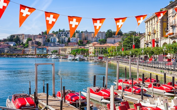 Lake Lugano waterfront with red pedal boats and Swiss flags, Switzerland.