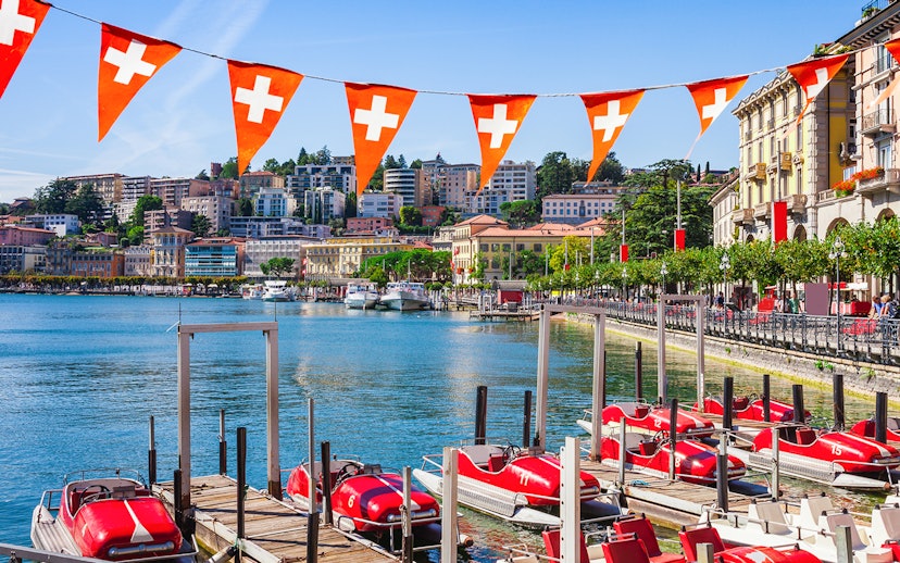 Lake Lugano waterfront with red pedal boats and Swiss flags, Switzerland.