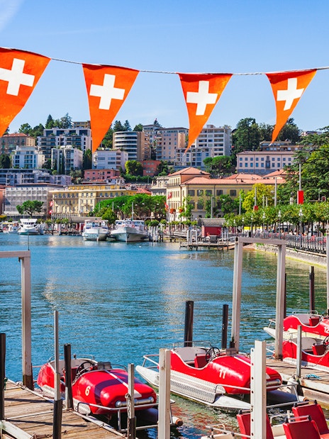 Lake Lugano waterfront with red pedal boats and Swiss flags, Switzerland.