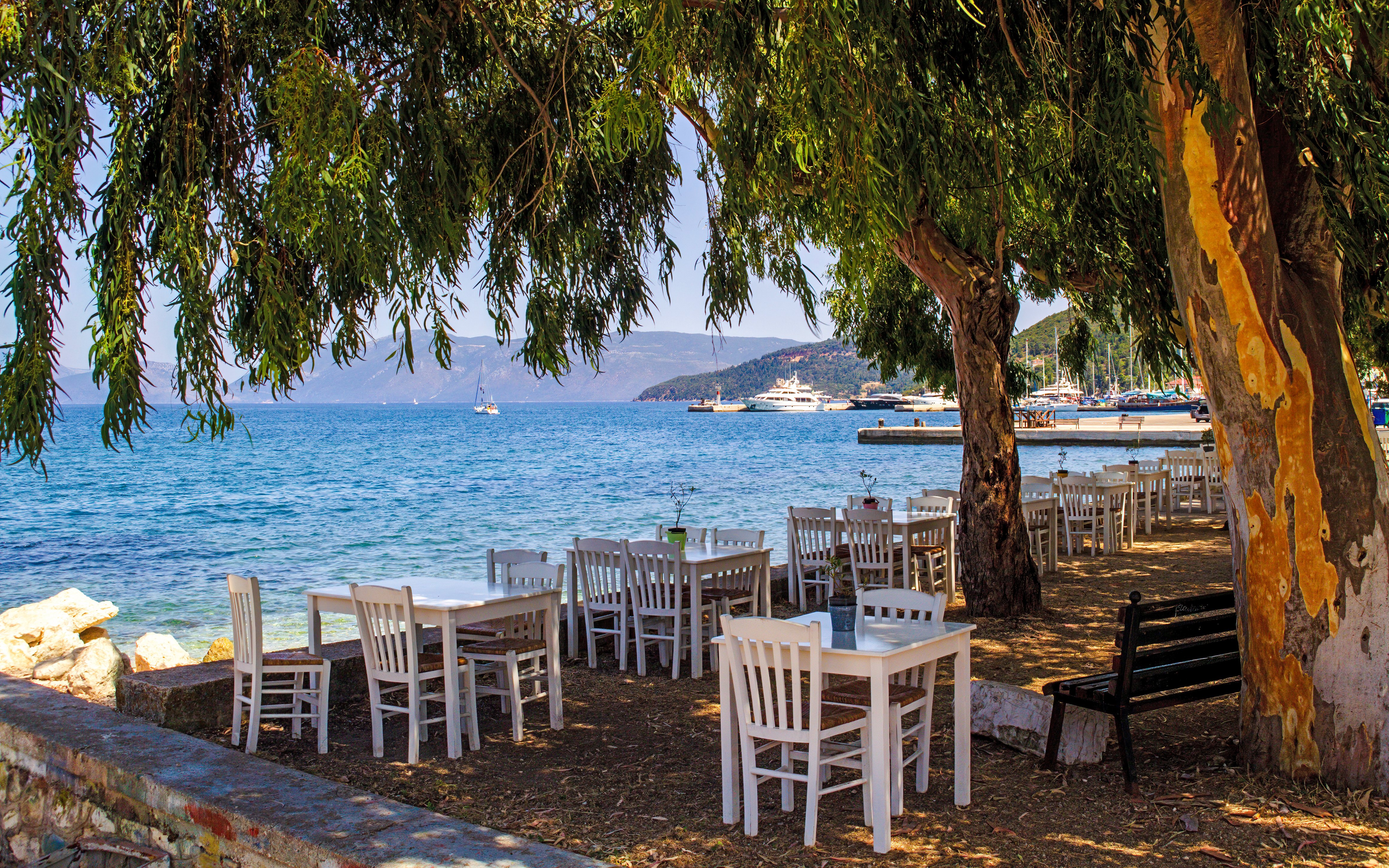 Tables under trees by the sea in Greece with boats in the distance.