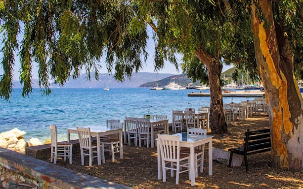 Tables under trees by the sea in Greece with boats in the distance.