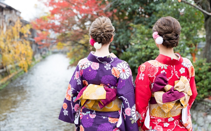 Women in colorful kimonos by a river in Kyoto during autumn.