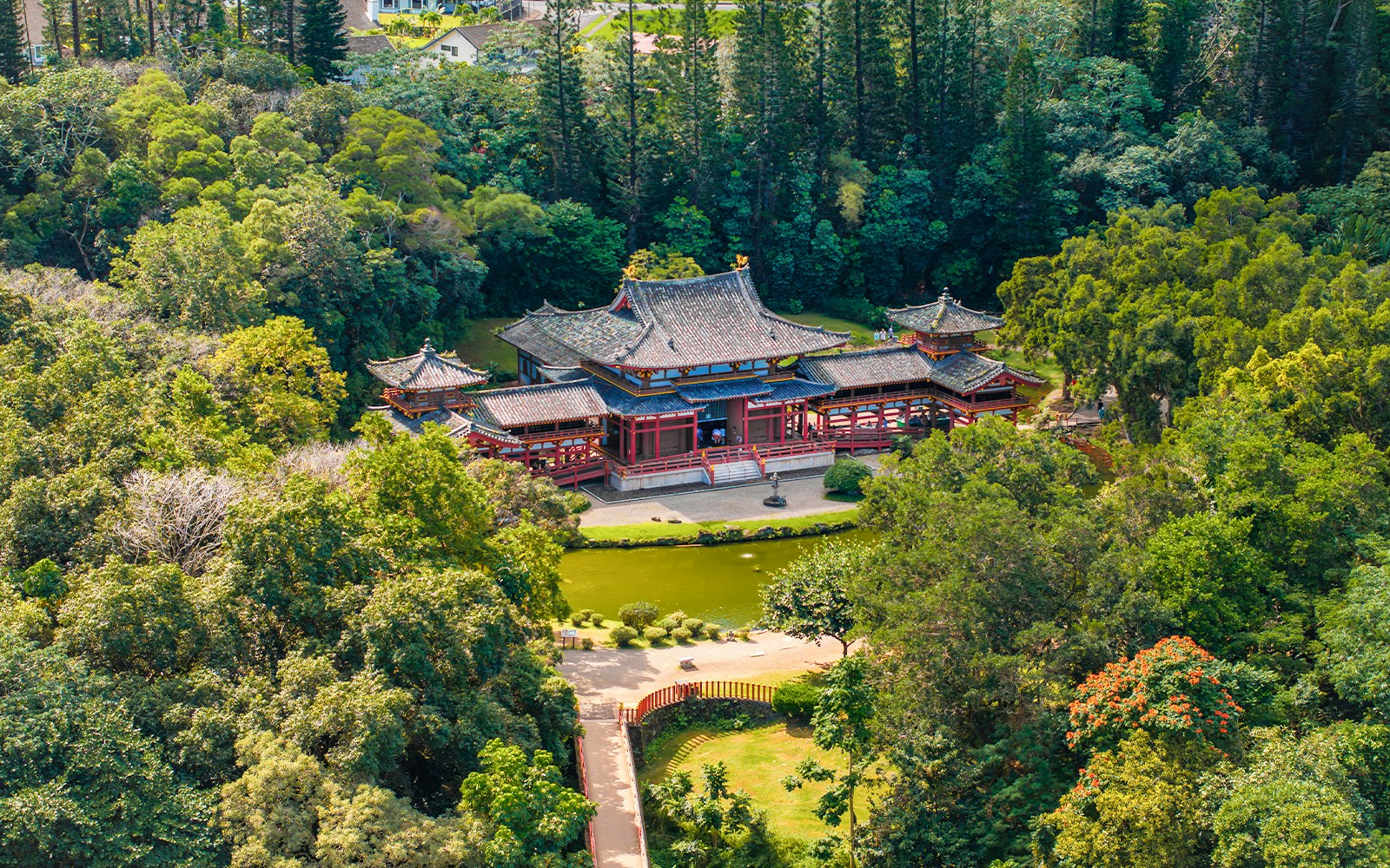 The Byodo In Temple in Oahu, Hawaii, features intricate architecture with red accents, set amidst a manicured garden, pond, and tall trees.
