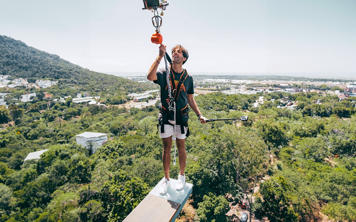Person standing on a plank with safety gear, high above a forested area, during Walk The Plank by AJ Hackett.