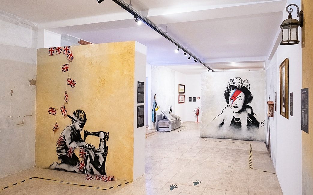 Banksy artwork in Lisbon museum featuring child with sewing machine and queen portrait.
