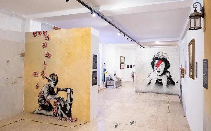 Banksy artwork in Lisbon museum featuring child with sewing machine and queen portrait.