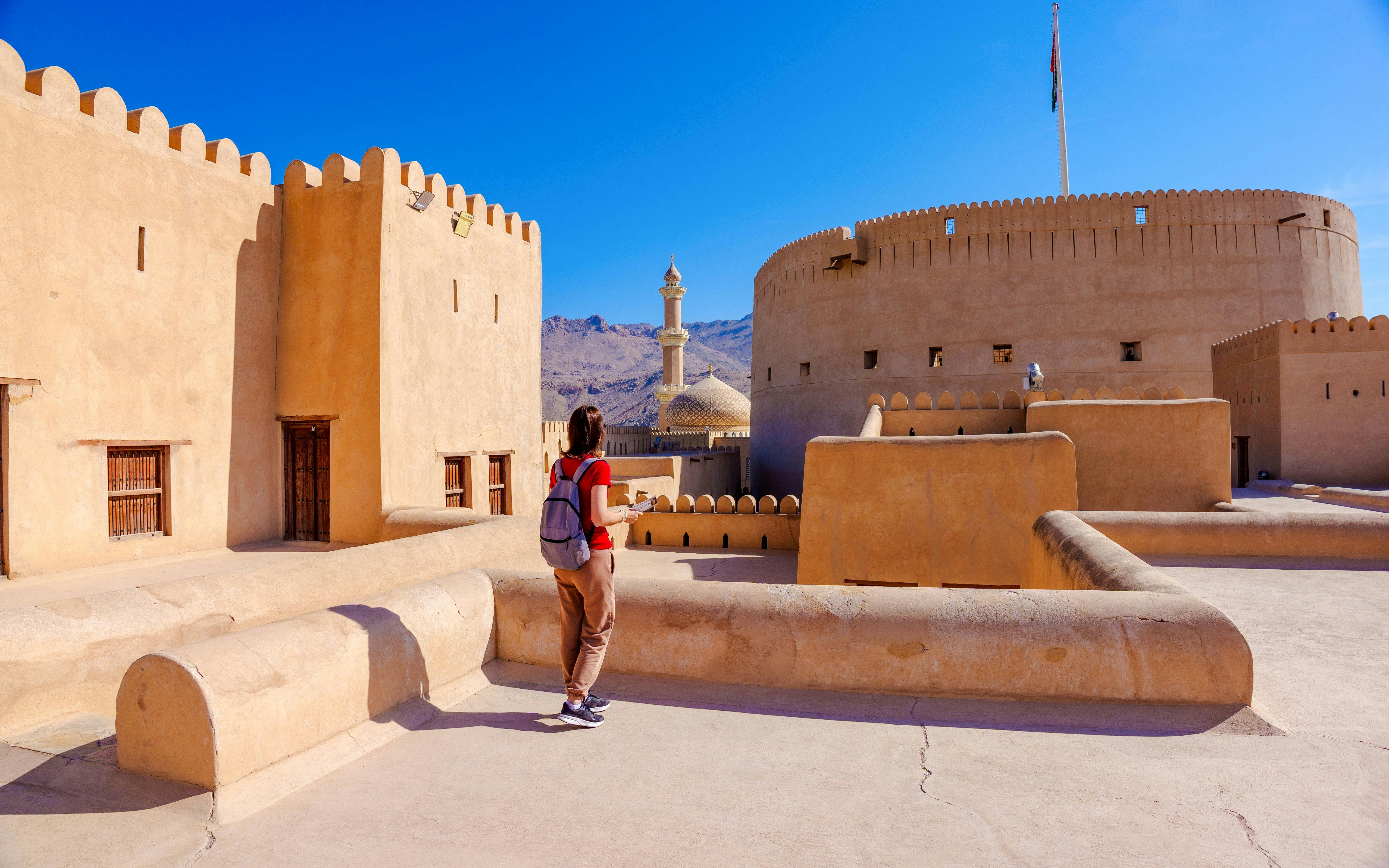 Visitor explores Nizwa Fort with minaret and mountains in background, Oman.