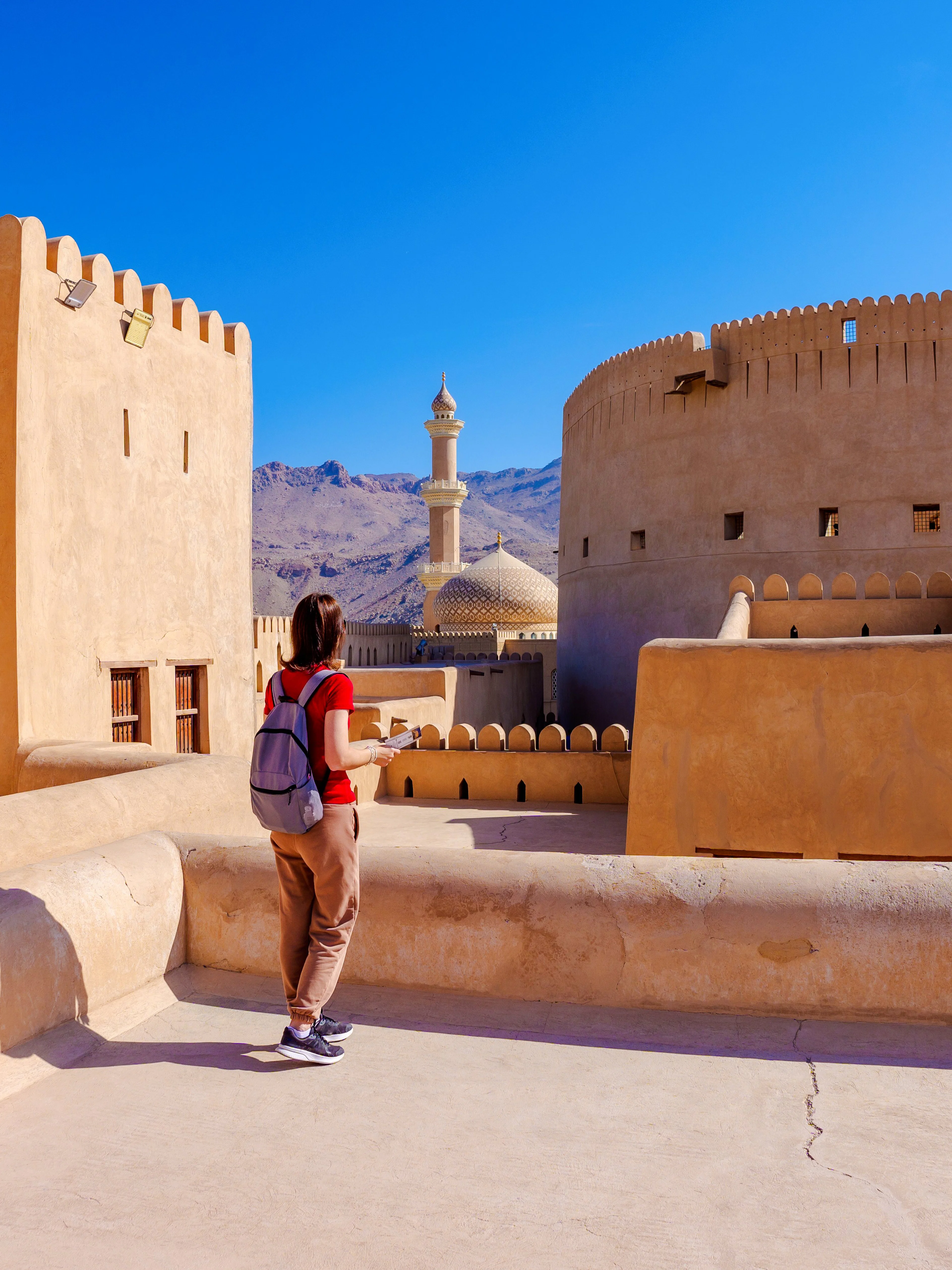 Visitor explores Nizwa Fort with minaret and mountains in background, Oman.