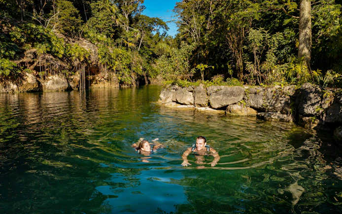 People swimming in a natural lagoon surrounded by lush greenery at Xcaret Park.