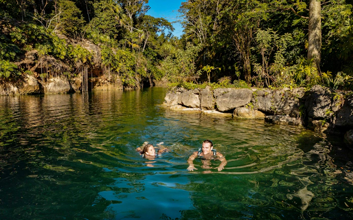 People swimming in a natural lagoon surrounded by lush greenery at Xcaret Park.