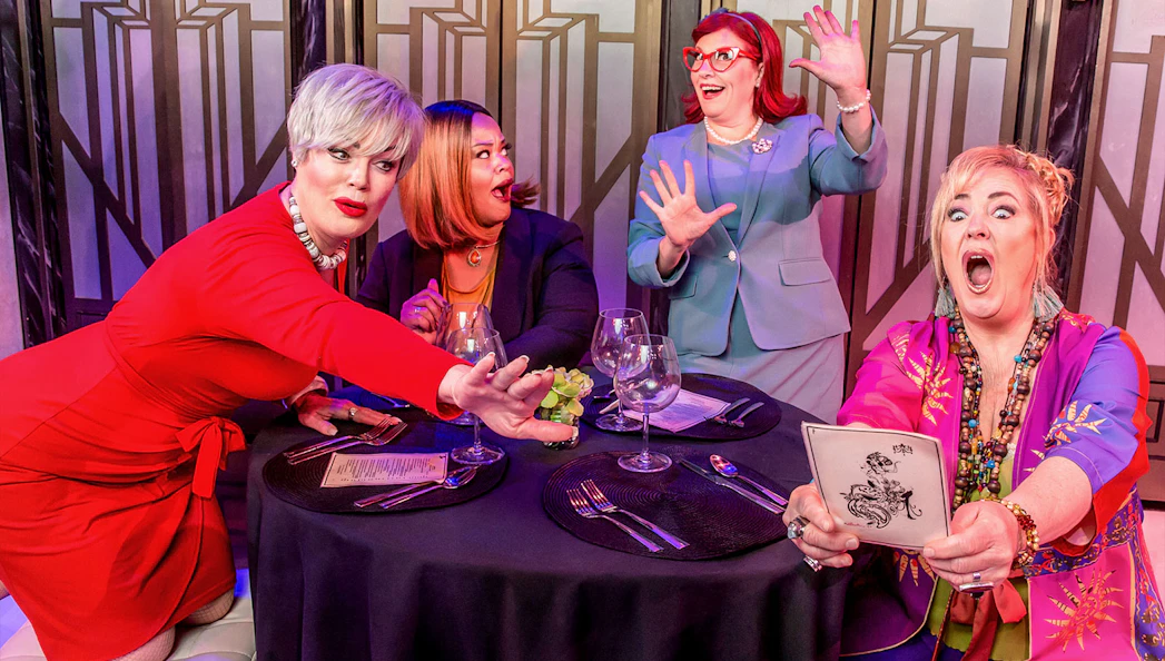 Women at a table reacting humorously during Menopause the Musical performance.