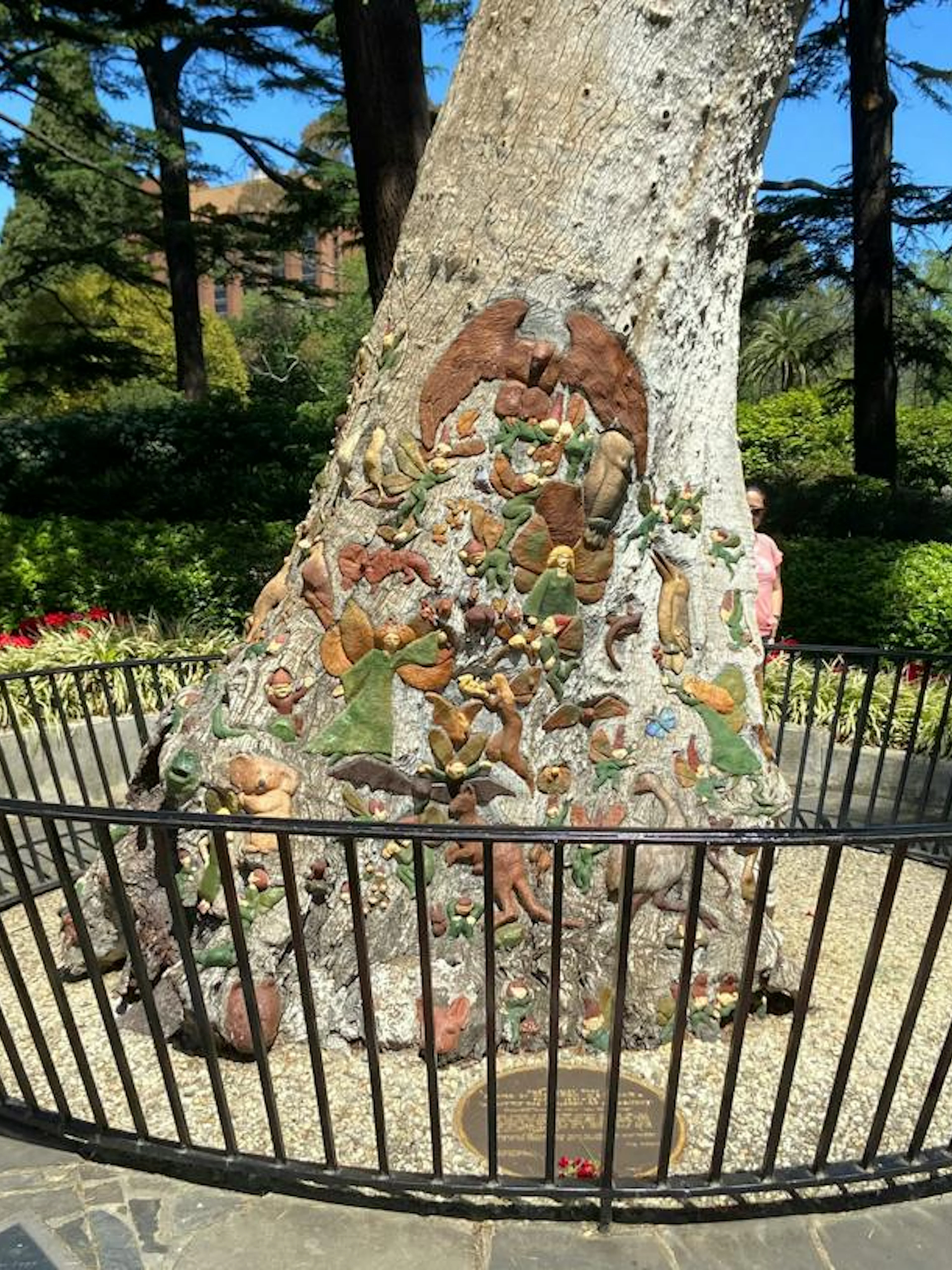 Tree trunk with intricate carvings in Fitzroy Gardens, Melbourne, surrounded by greenery and a black fence.
