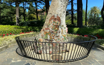 Tree trunk with intricate carvings in Fitzroy Gardens, Melbourne, surrounded by greenery and a black fence.