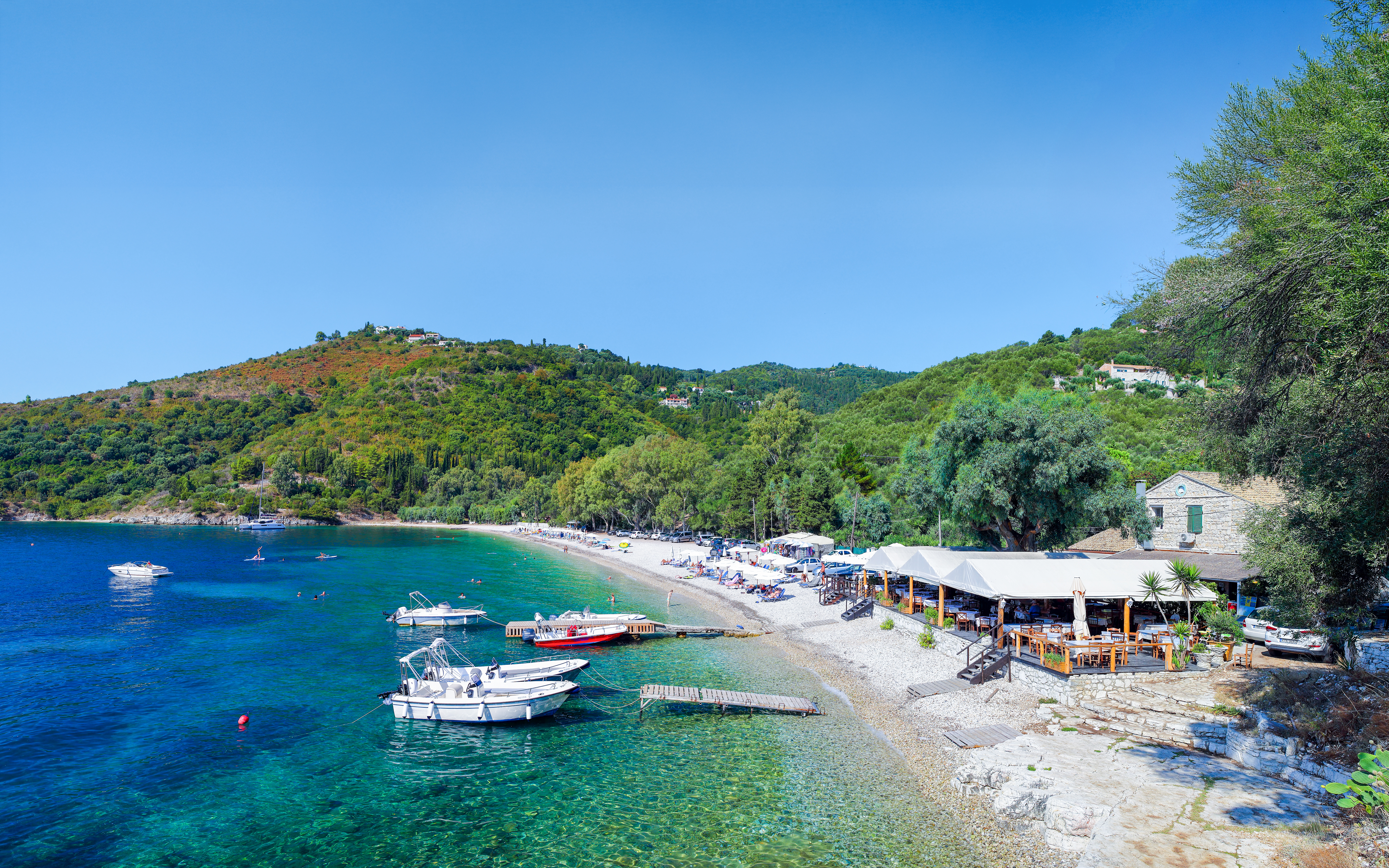 Boats anchored near Kerasia Beach, Corfu, Greece, with lush hills and a seaside restaurant.