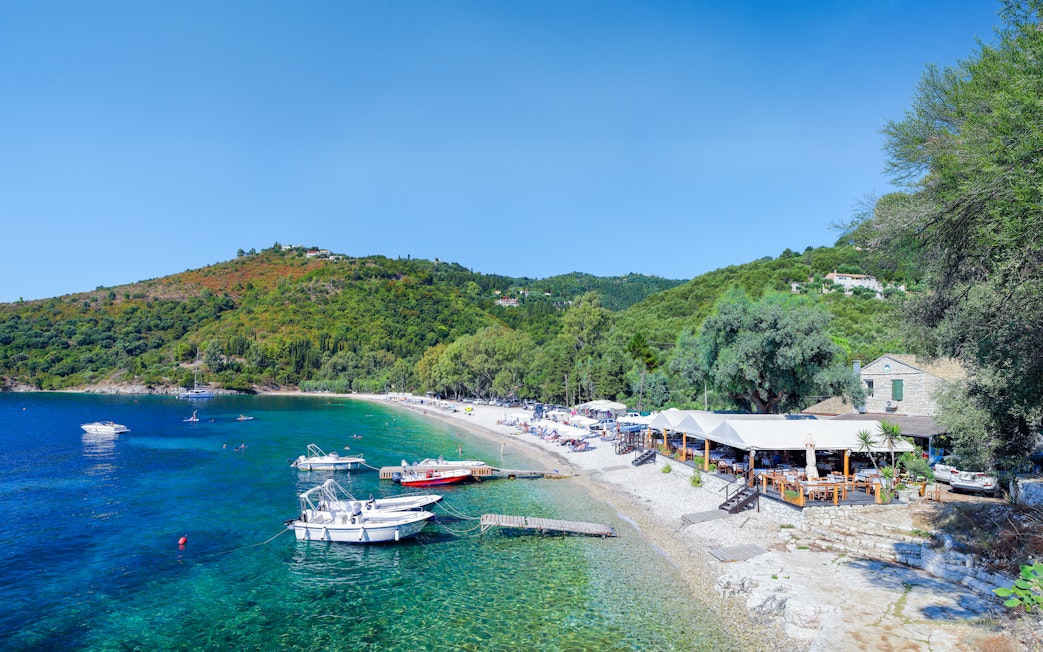 Boats anchored near Kerasia Beach, Corfu, Greece, with lush hills and a seaside restaurant.