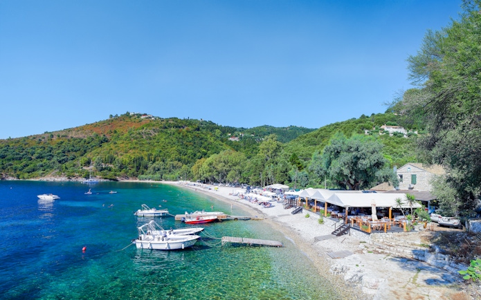 Boats anchored near Kerasia Beach, Corfu, Greece, with lush hills and a seaside restaurant.