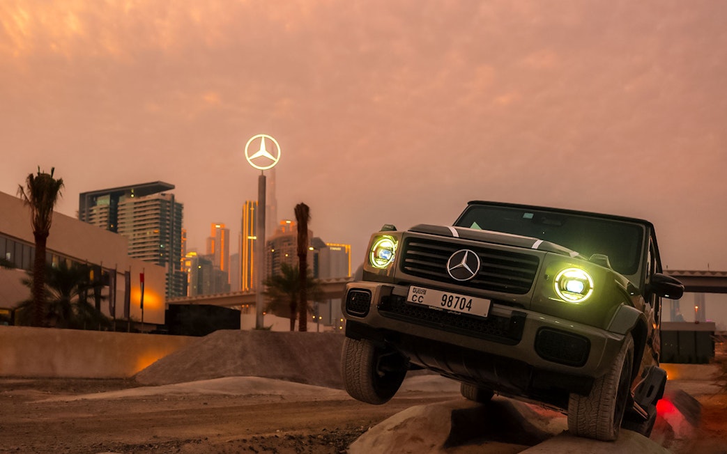 Mercedes-Benz SUV navigating terrain at sunset, Dubai skyline in background.