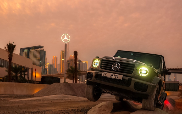 Mercedes-Benz SUV navigating terrain at sunset, Dubai skyline in background.