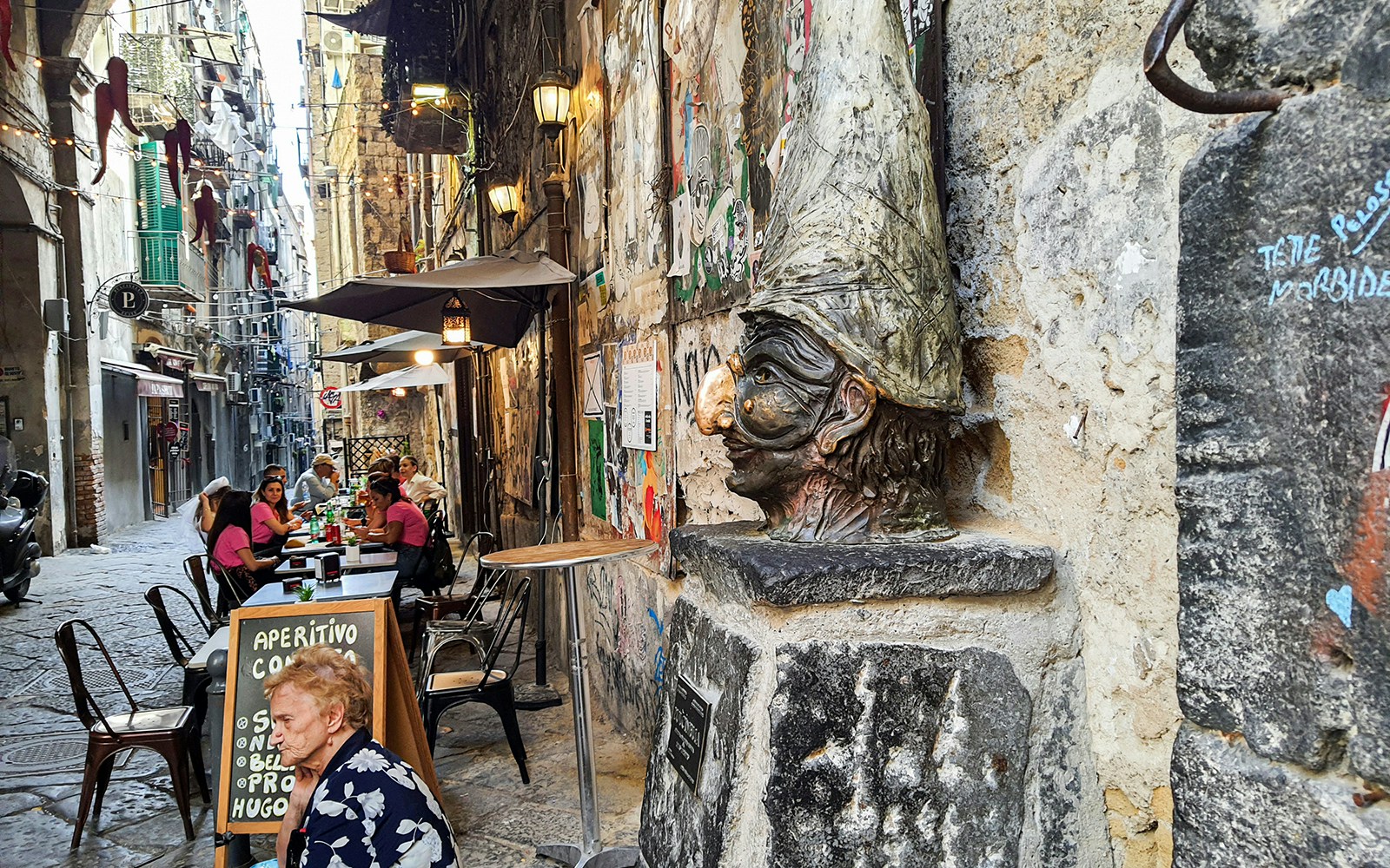 Bust of Pulcinella on a narrow street with outdoor dining in Naples.