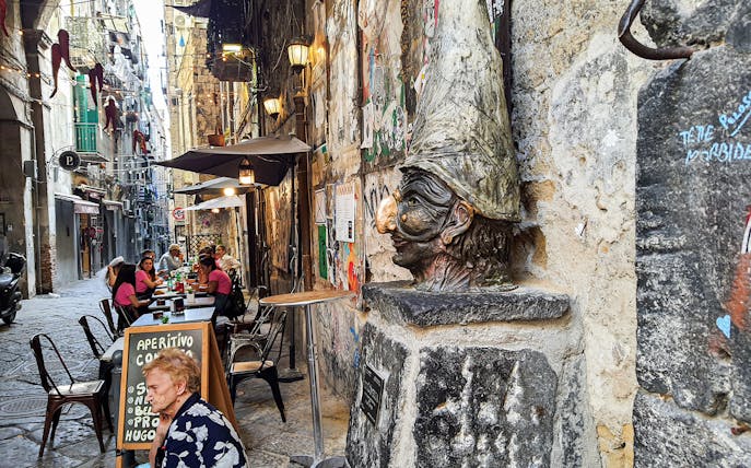 Bust of Pulcinella on a narrow street with outdoor dining in Naples.