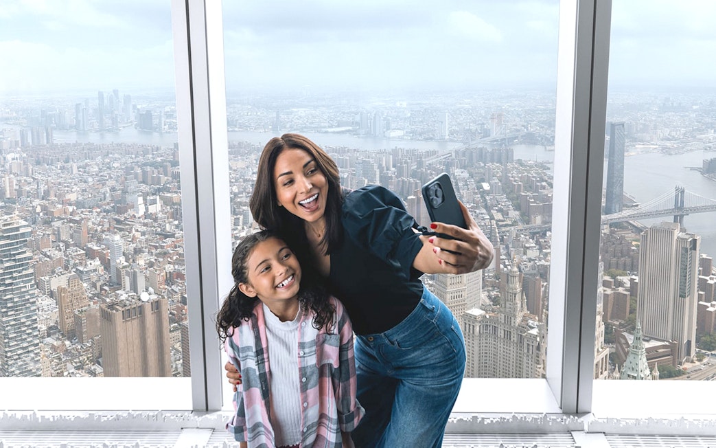 Visitors taking a selfie at One World Observatory with New York City skyline in the background.