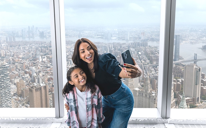 Visitors taking a selfie at One World Observatory with New York City skyline in the background.