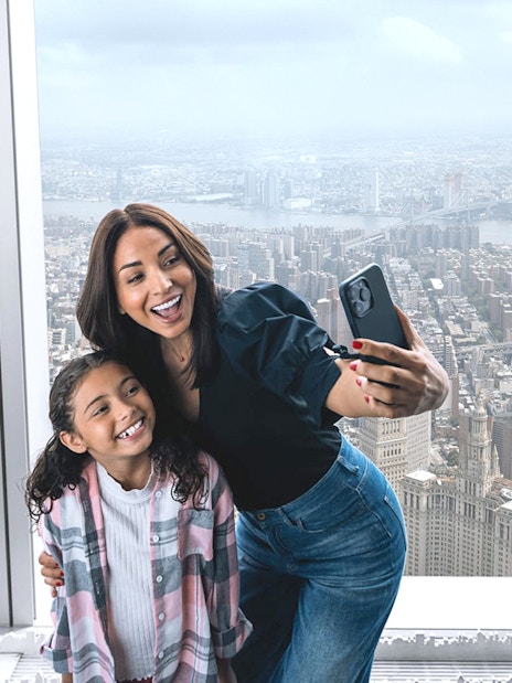 Visitors taking a selfie at One World Observatory with New York City skyline in the background.