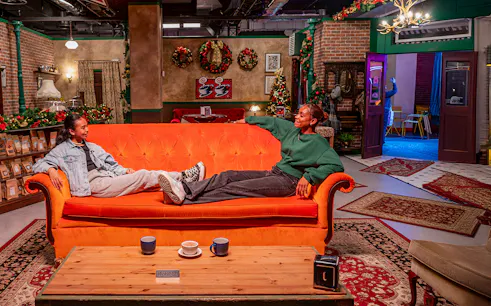 Visitors relaxing on the iconic orange couch at The FRIENDS™ Experience, surrounded by festive decor.