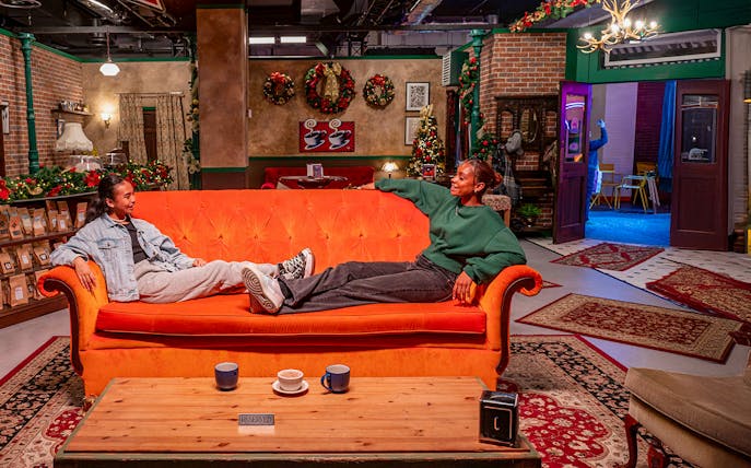Visitors relaxing on the iconic orange couch at The FRIENDS™ Experience, surrounded by festive decor.
