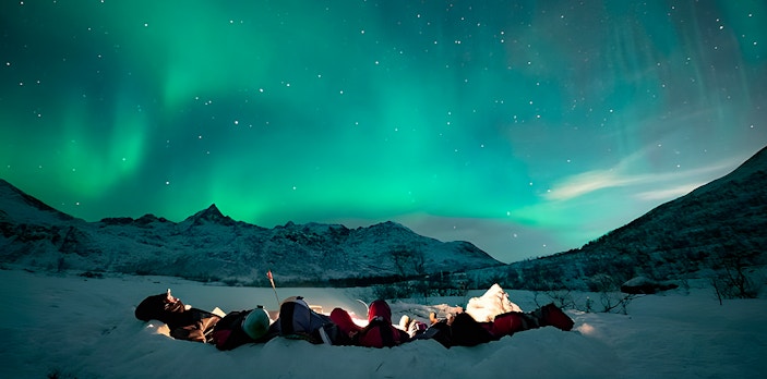 People lying on snow in Tromso watching Northern Lights display.