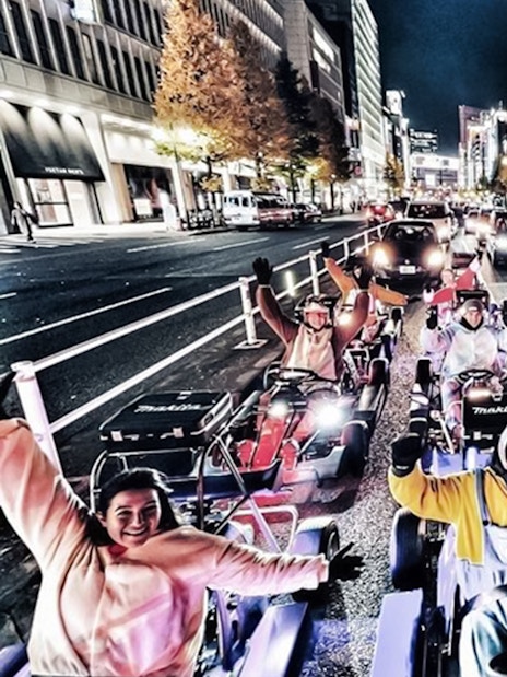Group in costumes on go-karts driving through a city street at night.