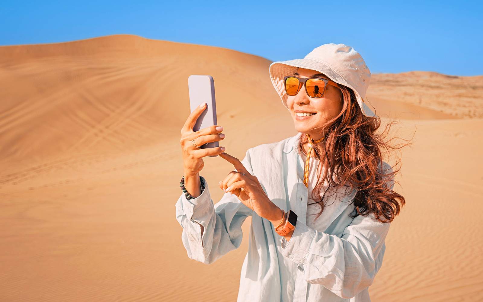 Woman in desert wearing sunglasses and hat, taking a photo with smartphone.
