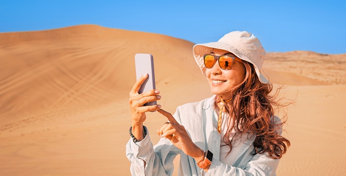 Woman in desert wearing sunglasses and hat, taking a photo with smartphone.