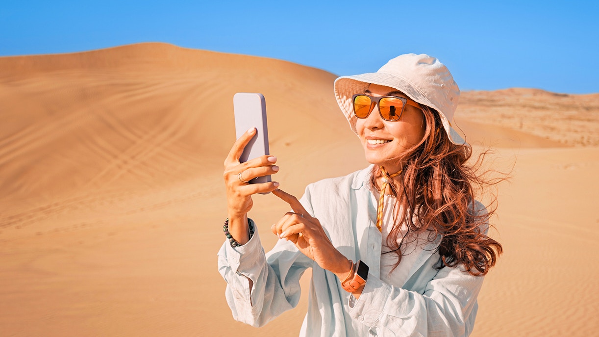 Woman in desert wearing sunglasses and hat, taking a photo with smartphone.