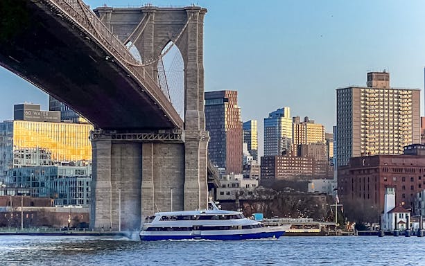 Cruise boat passing under Brooklyn Bridge with Manhattan skyline in view.