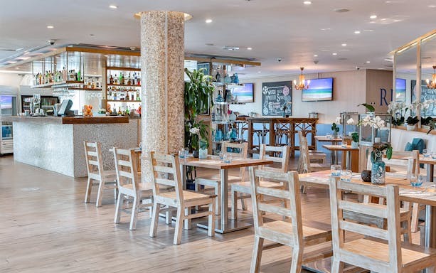 Riva Beach Club dining area with wooden tables and chairs, bar in the background.