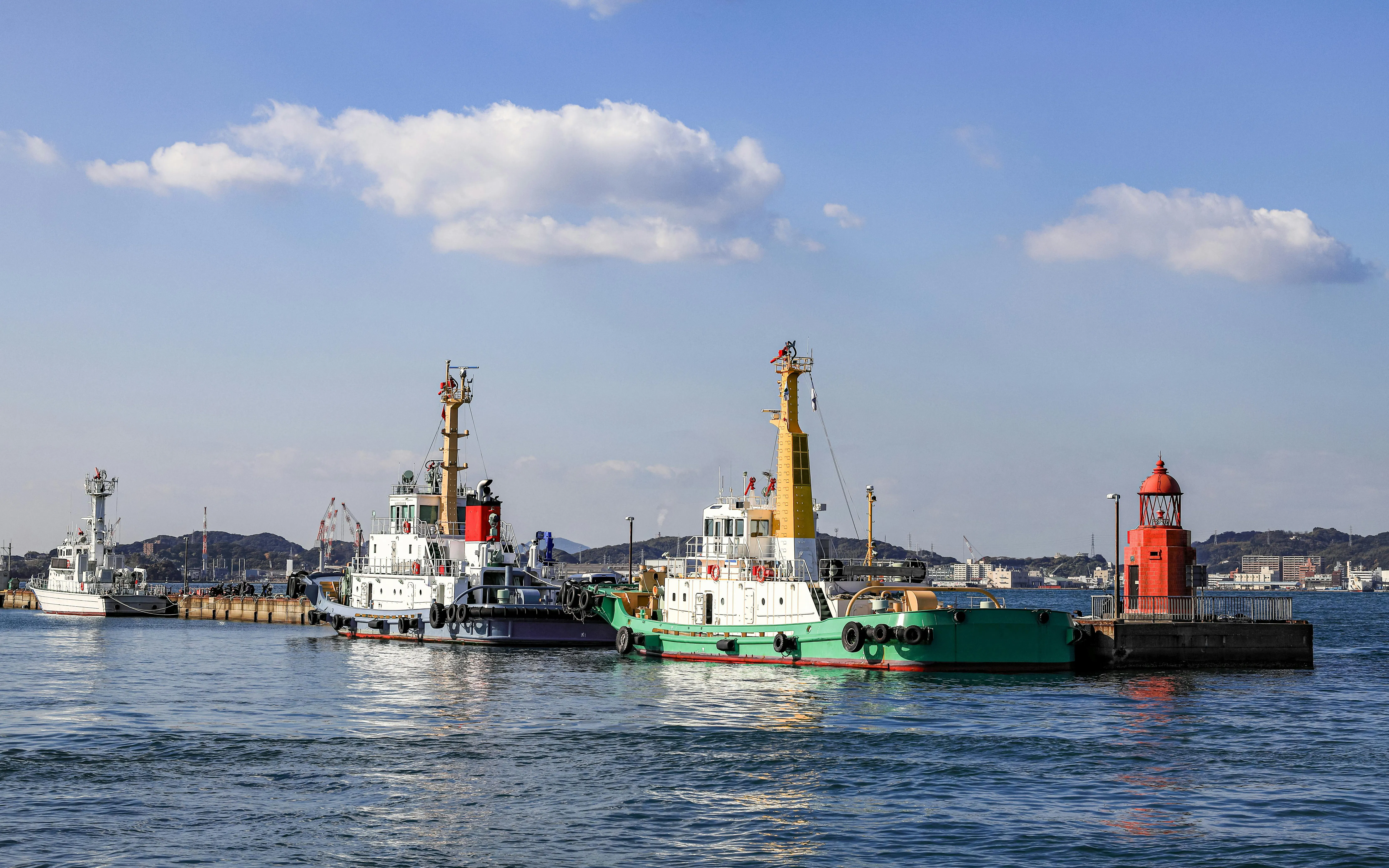 Tug boats docked at Moji Port in Kitakyushu, Japan with a red lighthouse nearby.