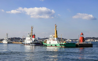 Tug boats docked at Moji Port in Kitakyushu, Japan with a red lighthouse nearby.