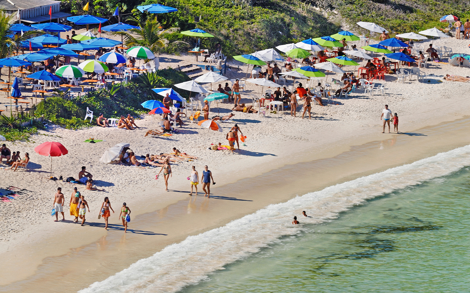 Beachgoers relax under umbrellas on Forno Beach, Arraial do Cabo, Brazil, with clear waters and lush forest.