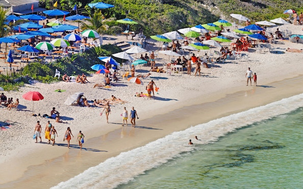 Beachgoers relax under umbrellas on Forno Beach, Arraial do Cabo, Brazil, with clear waters and lush forest.