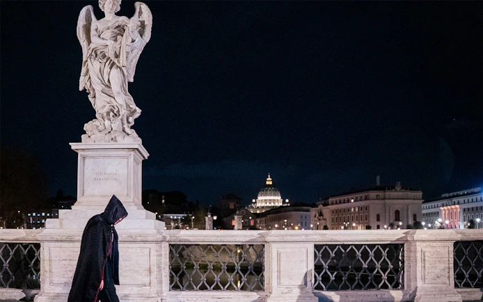 Cloaked figure walking by angel statue on Rome's bridge at night.