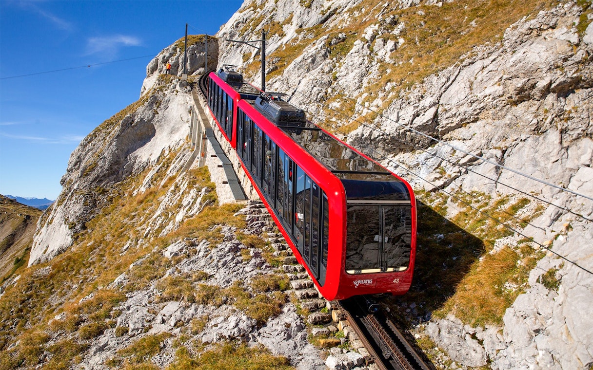 Red cogwheel train ascending Mount Pilatus, Switzerland, with rocky landscape.